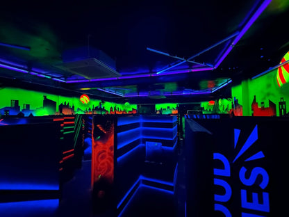 A neon-lit laser tag arena with glowing blue and green walls, dark maze-like structures, and a cityscape mural.