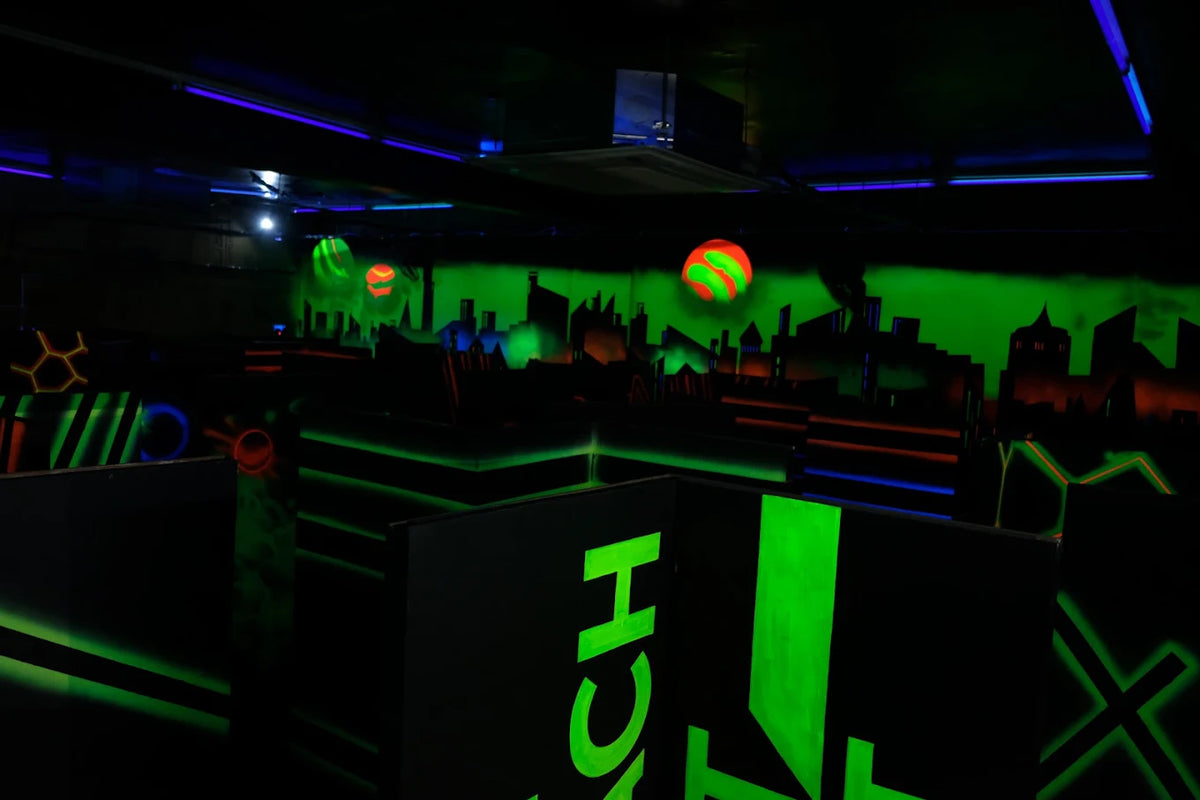A dark laser tag arena with neon green and blue lights, featuring a cityscape mural and glowing balls.