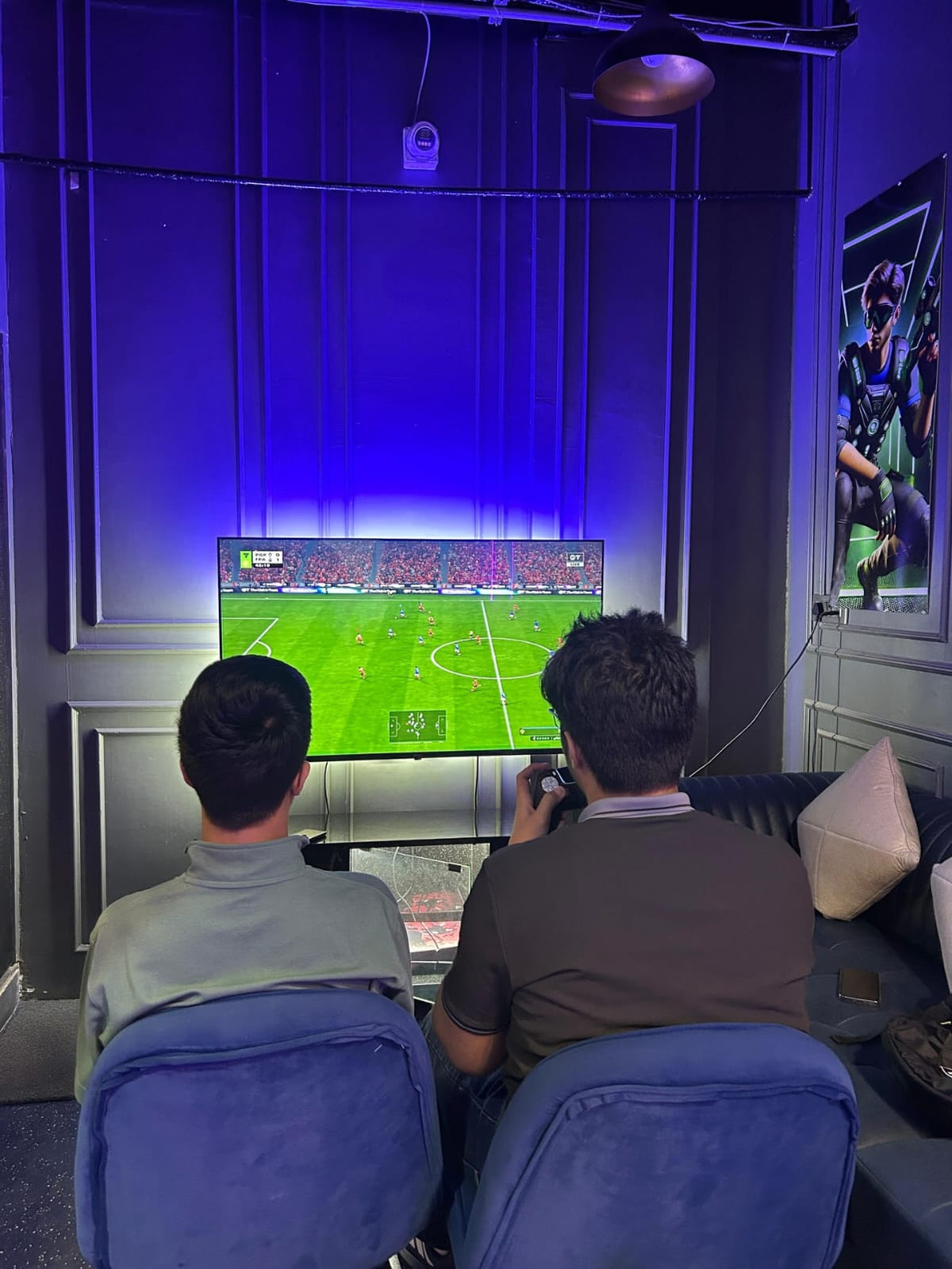 Two young men play a soccer video game in a room with blue lighting and a large screen.