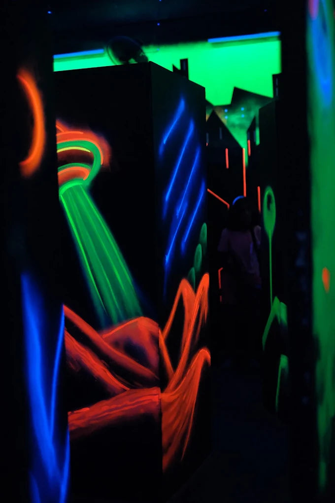 A person walks through a dark laser tag arena with neon green, blue, and orange lights.