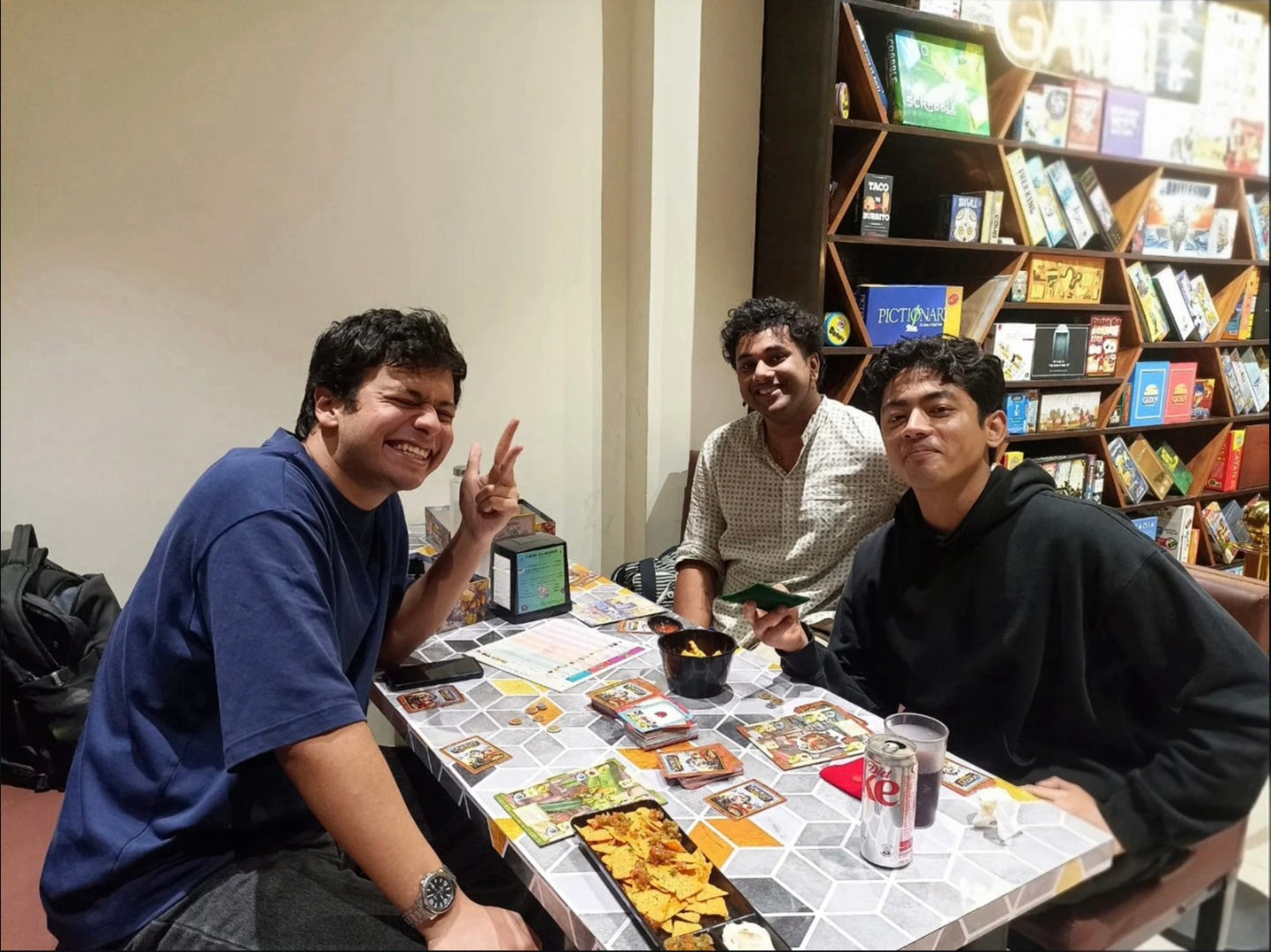 Three young men play a board game at a table filled with game cards and snacks, with shelves of games in the background.