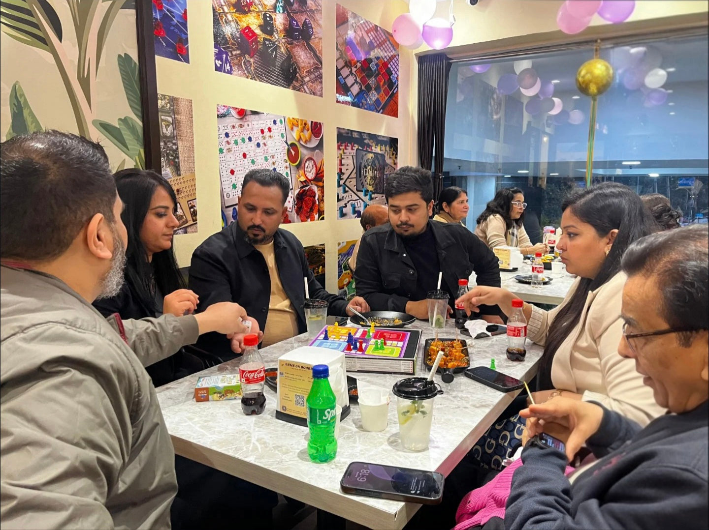 A group of adults plays a board game at a table in a restaurant, with food and drinks present.