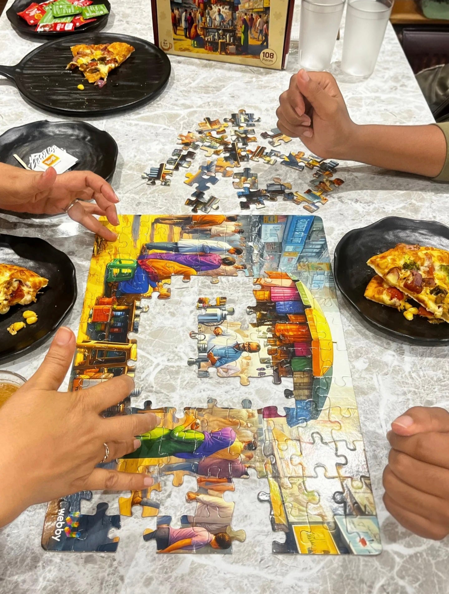 People are assembling a puzzle of a colorful street scene while eating pizza.