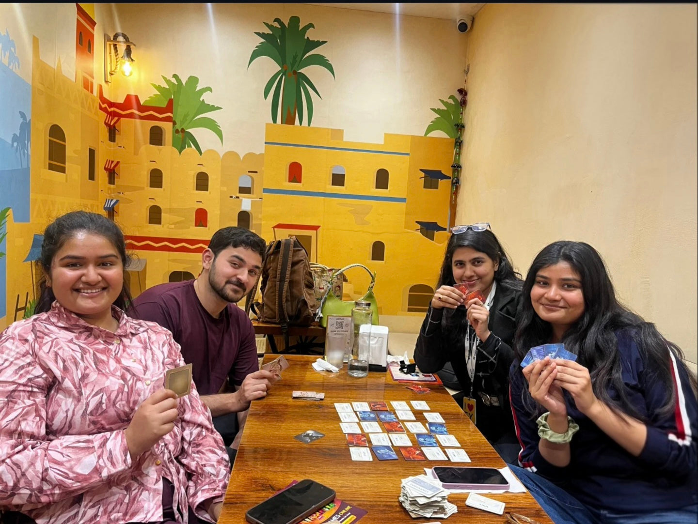 Four young adults play a card game at a wooden table in front of a colorful mural.