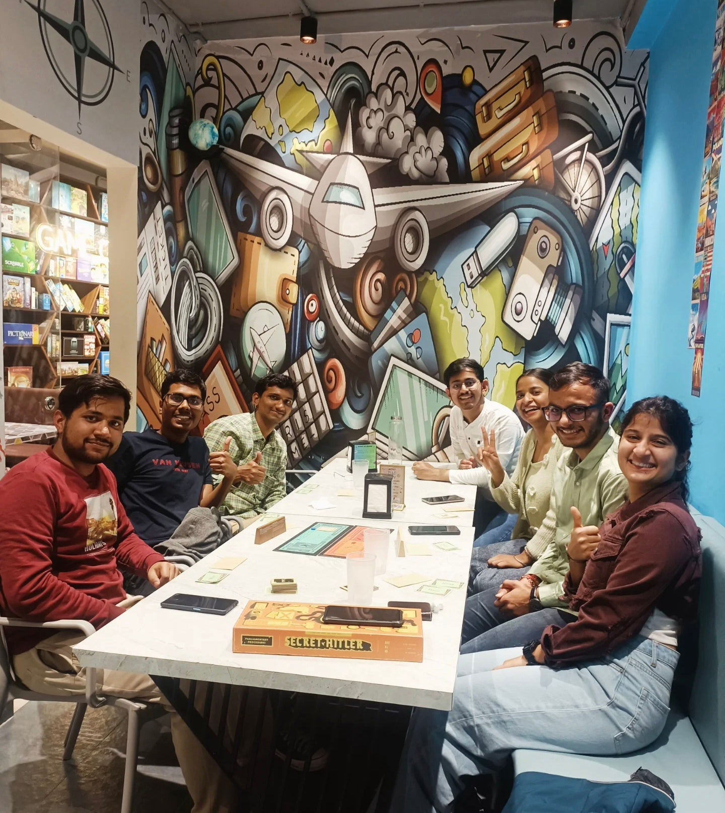 Young adults play a board game at a table in a cafe with a travel-themed mural.