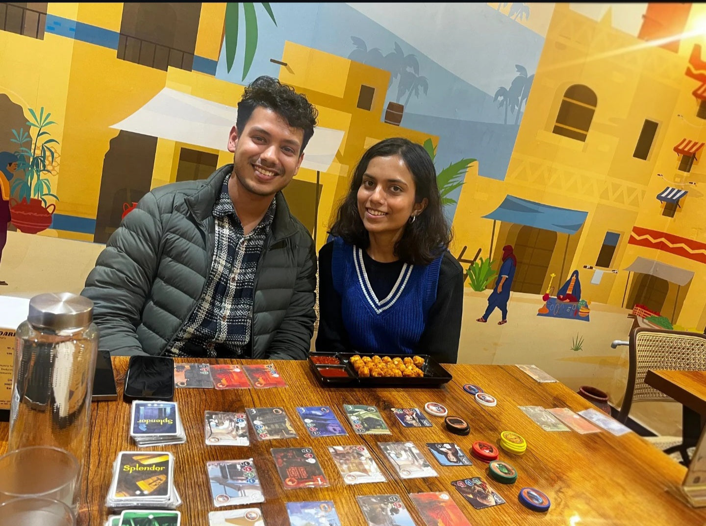 Two people sitting at a table with card games and snacks in front of a colorful mural.
