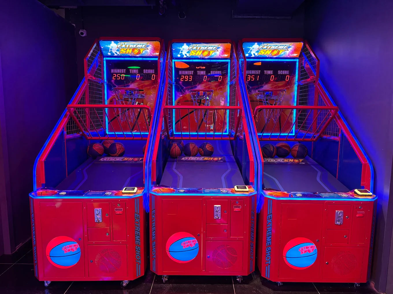 Three red arcade basketball shooting games with blue neon lights, set up side-by-side in an entertainment venue.