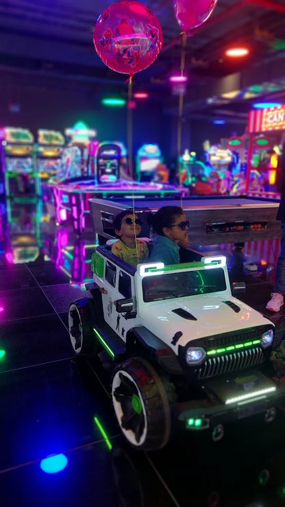 Two young children in sunglasses ride in a toy jeep at a brightly lit arcade.