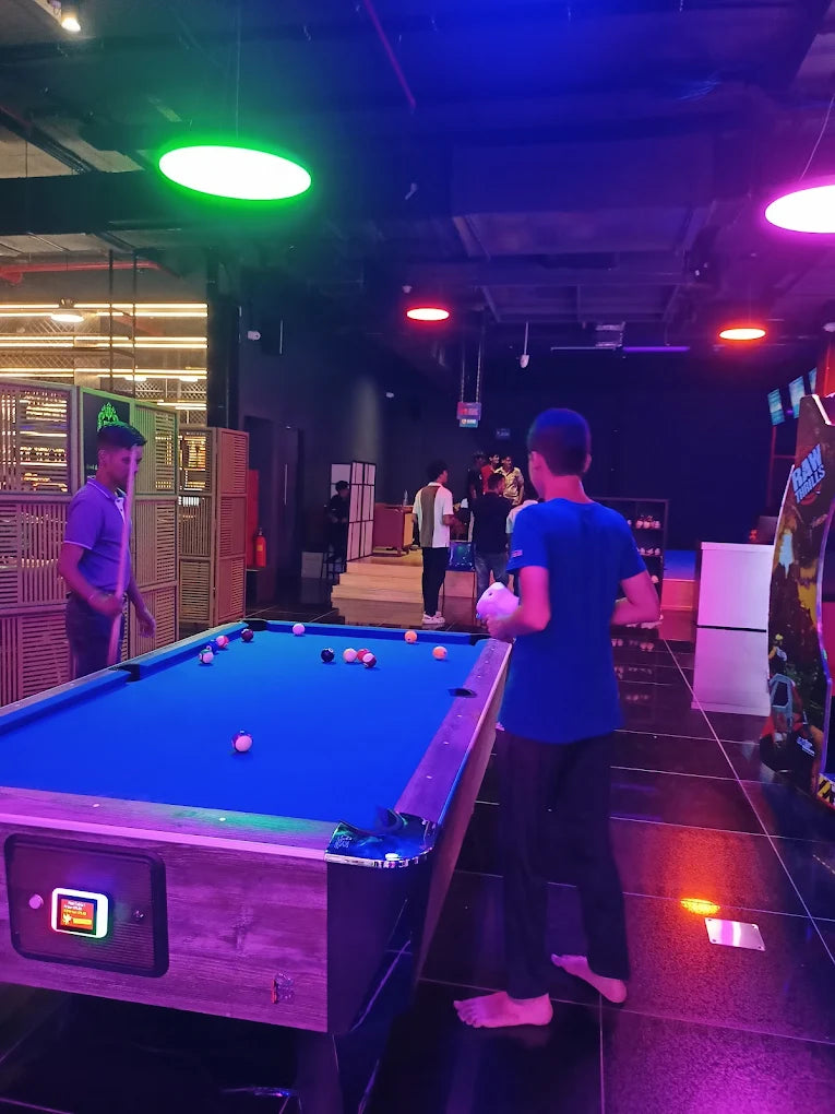 Two young men play pool in a dimly lit arcade with neon lights.
