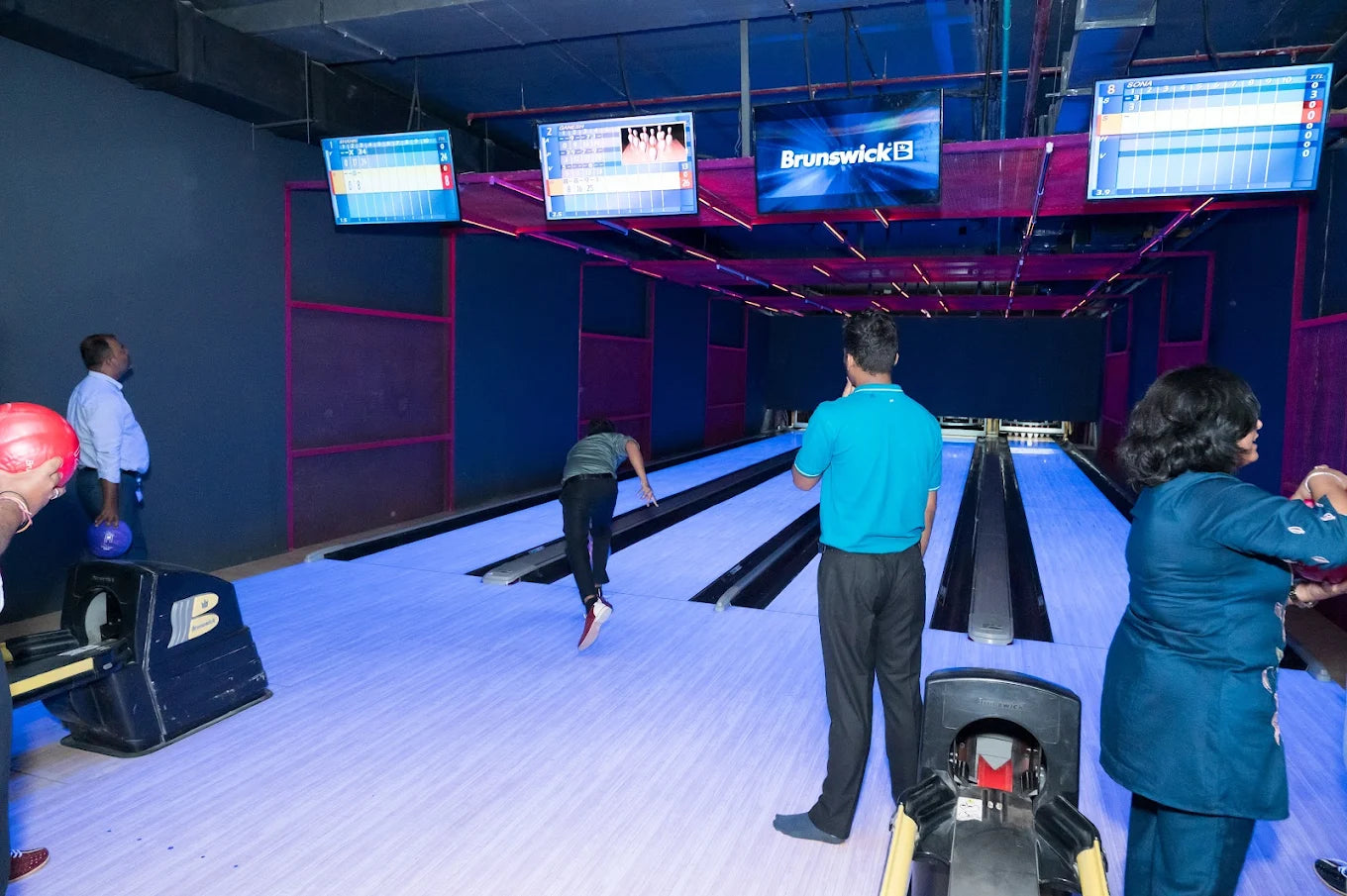 Adults play bowling in a dimly lit alley with glowing lanes and scoreboards.