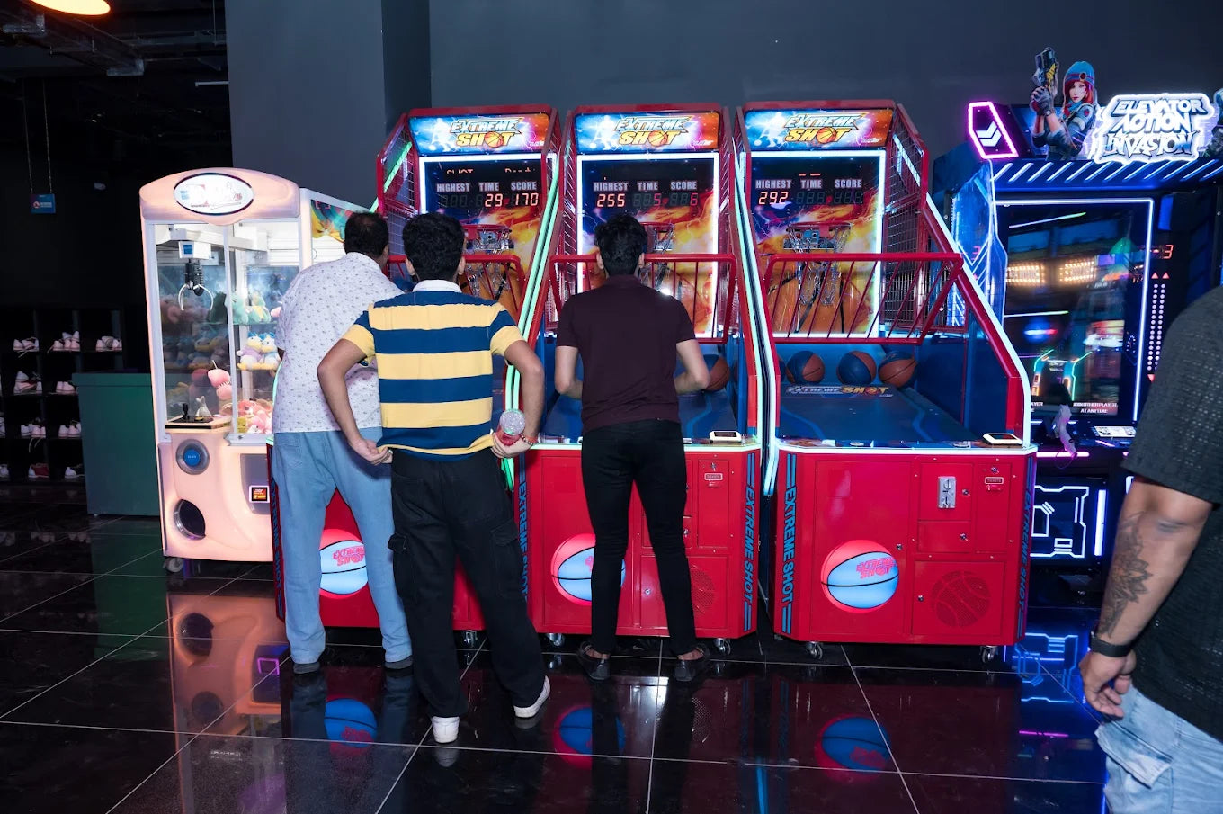Young adults play basketball arcade games in a dimly lit entertainment center.