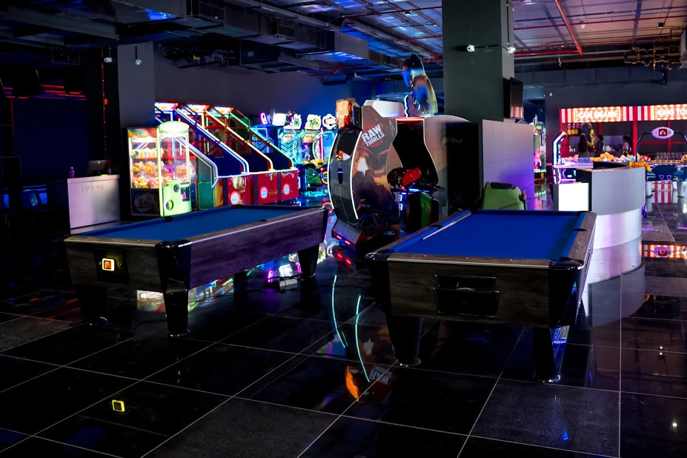 Two pool tables in an arcade with glowing game machines.