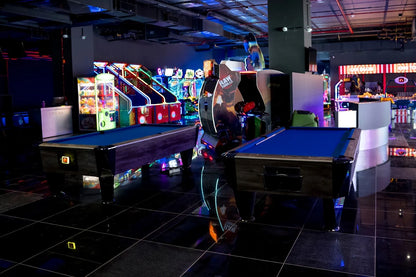 Two pool tables in an arcade with glowing game machines.
