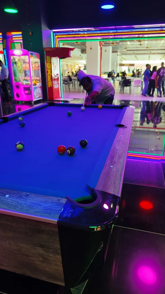 A person plays pool in a brightly lit arcade with neon lights. Several colorful billiard balls are scattered on the blue felt table.