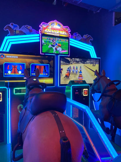 Two people ride simulated horses in a brightly lit arcade, playing a horse racing game.
