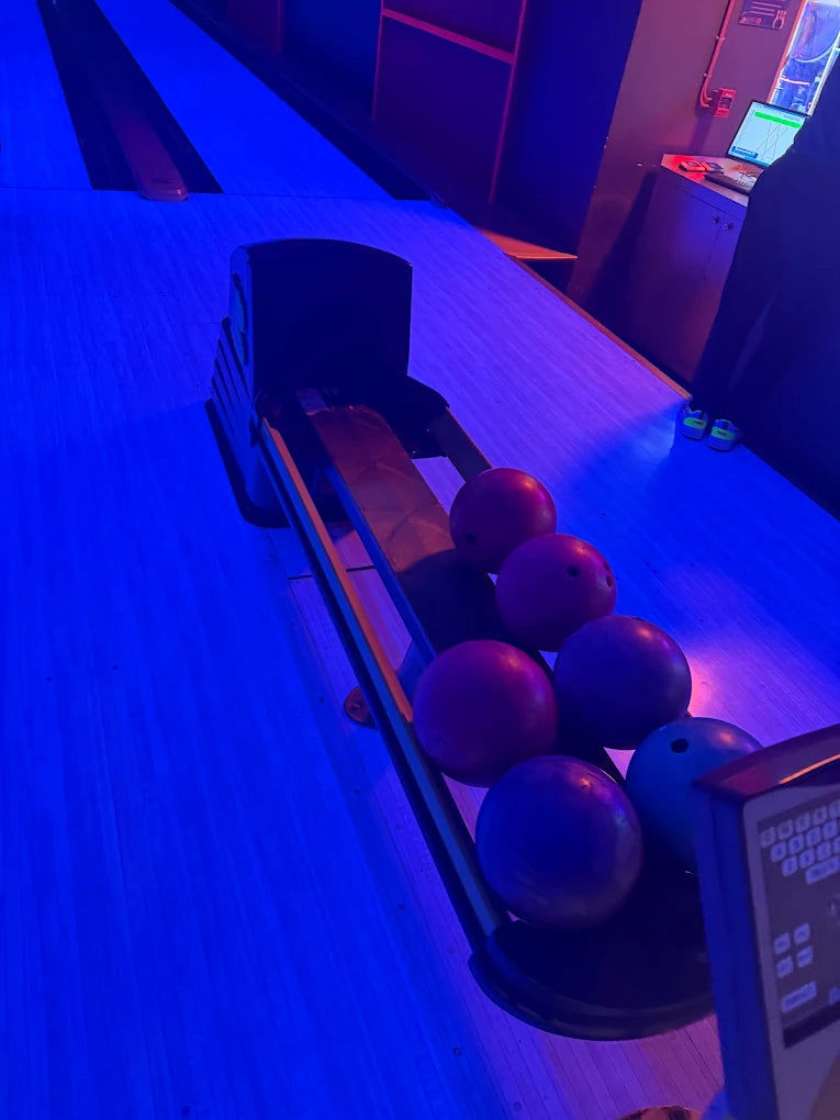 Bowling balls rest on a lane under blue lights, with a scoring console visible.