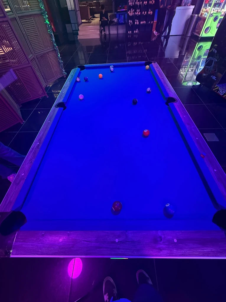 A group of young adults play pool in a dimly lit arcade with neon lights.