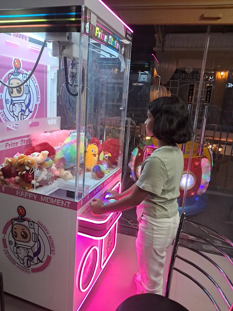 A young child plays a claw machine filled with stuffed animals, illuminated by pink neon lights.