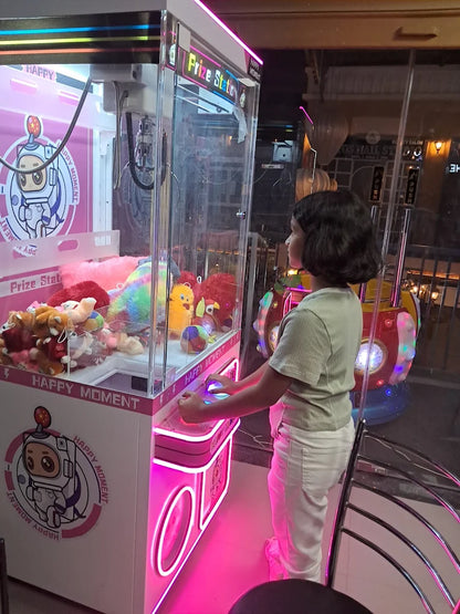 A young child plays a claw machine filled with stuffed animals, illuminated by pink neon lights.