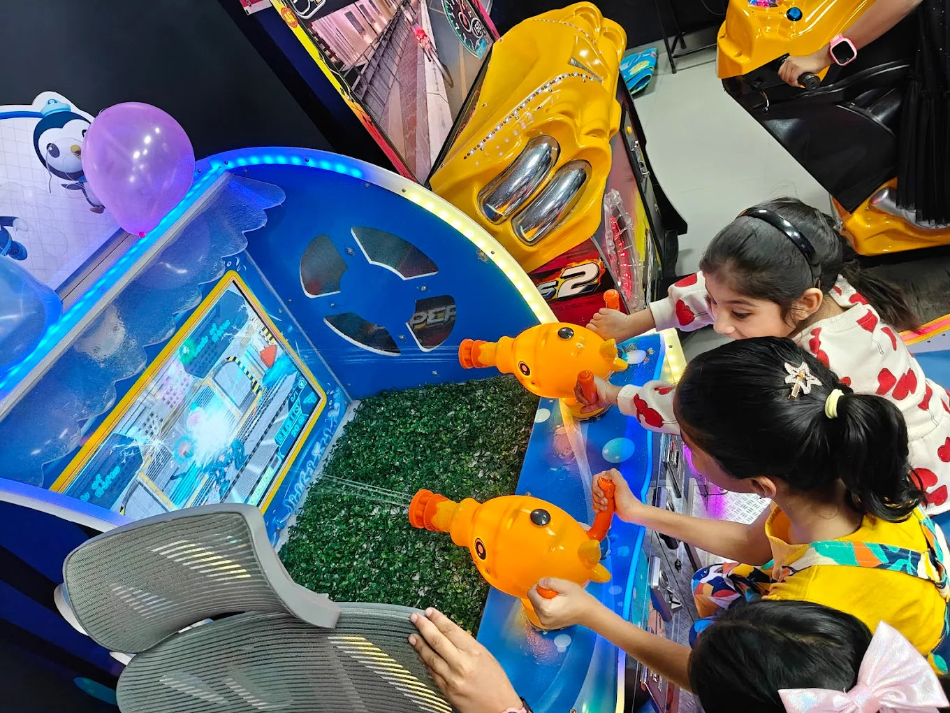 Young girls play a water gun arcade game, aiming at a screen with orange fish-shaped guns.