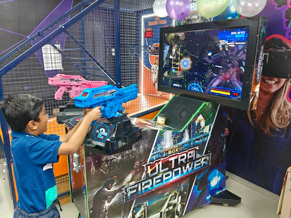 A young boy plays an arcade game with a blue gun, while a woman in VR goggles watches.