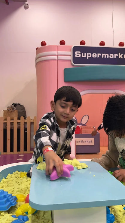 A young boy plays with kinetic sand in a supermarket-themed play area. He is holding a purple star-shaped toy.