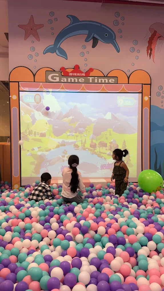 Children play in a ball pit in front of a large screen displaying a video game.