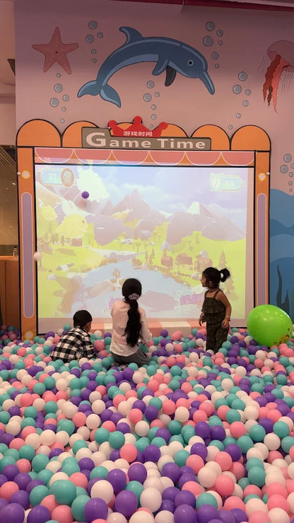 Children play in a ball pit in front of a large screen displaying a video game.