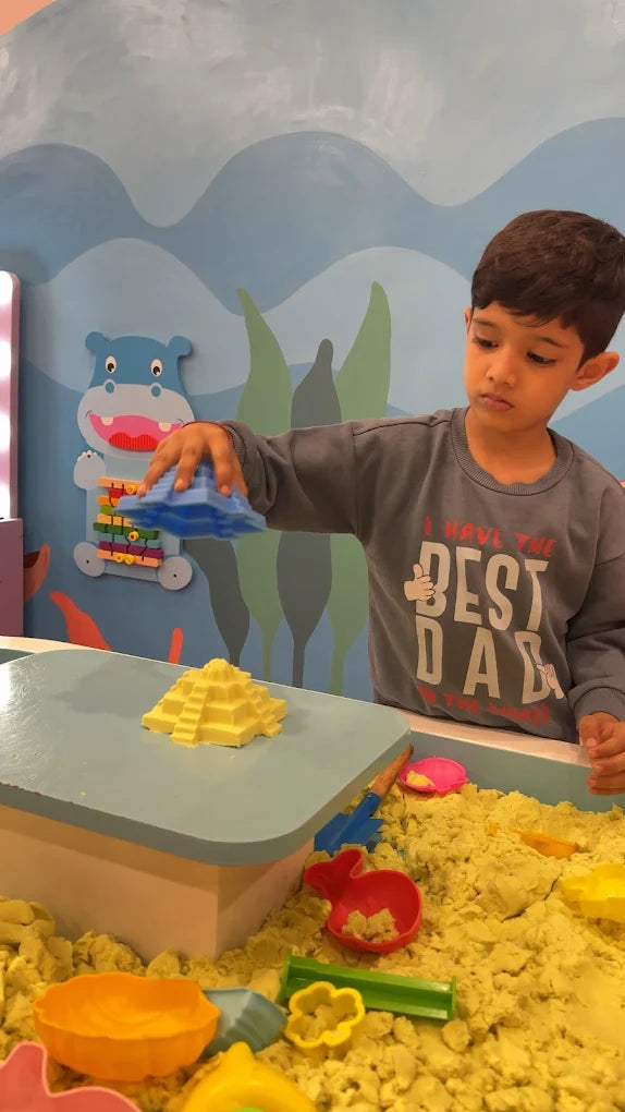 A young boy plays with kinetic sand, building with a pyramid mold in a colorful play area.