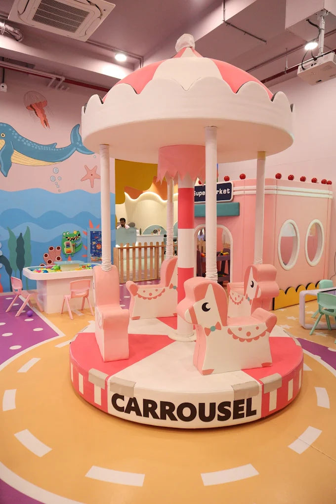 A pink and white carousel with horse seats in a brightly colored indoor play area for children.