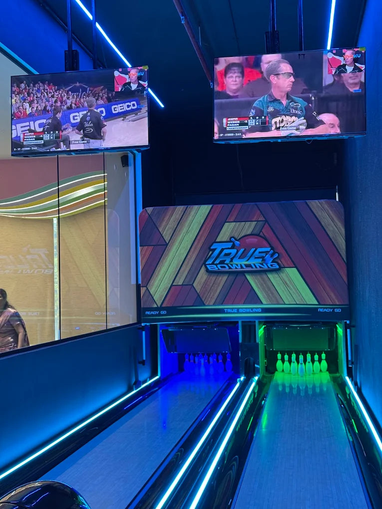 Two young adults are bowling in a brightly lit alley with screens showing a bowling competition.