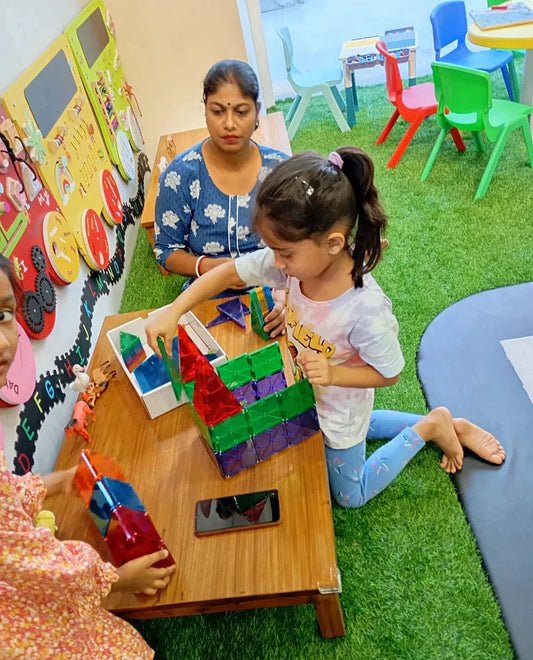 A woman and two young children play with magnetic building blocks at a wooden table in a classroom.