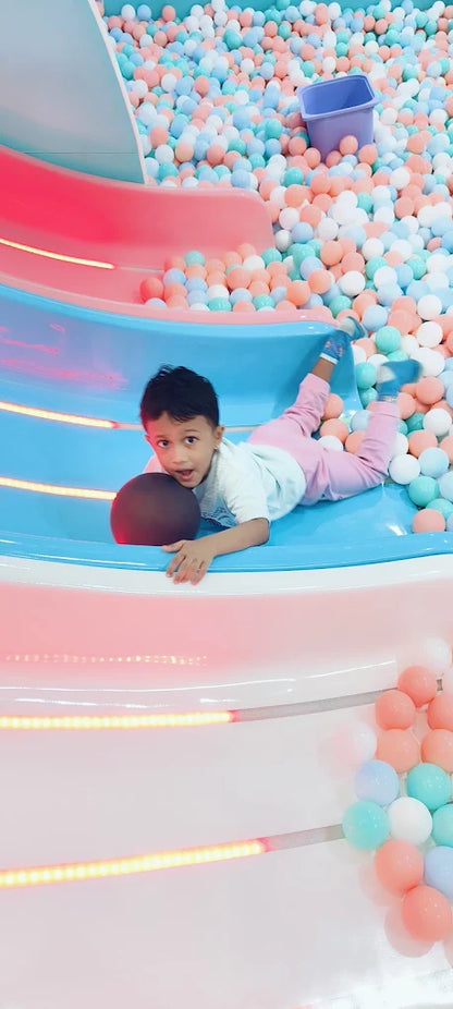 A young child slides down a blue and pink slide into a ball pit filled with pastel-colored balls.