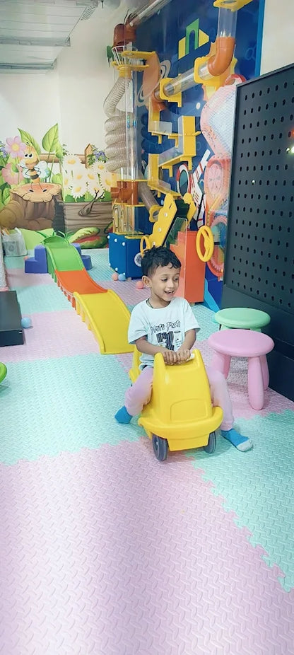A young boy rides a yellow toy car in a colorful indoor play area with slides and tubes.