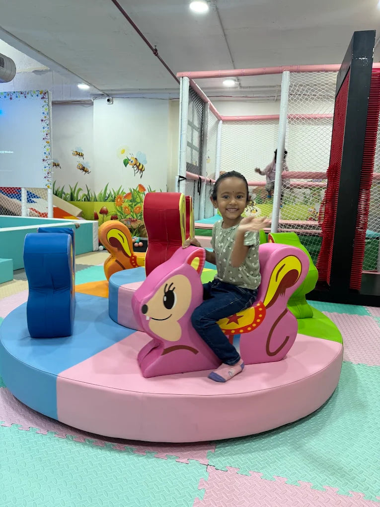 A young girl rides a pink squirrel carousel in a brightly colored indoor play area.