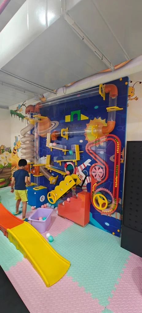 A young boy plays with a ball maze toy on a colorful mat in a playroom. The maze features tubes, gears, and slides.