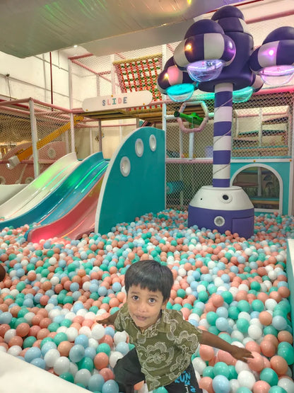 A young boy plays in a ball pit at an indoor playground with slides and colorful decor.