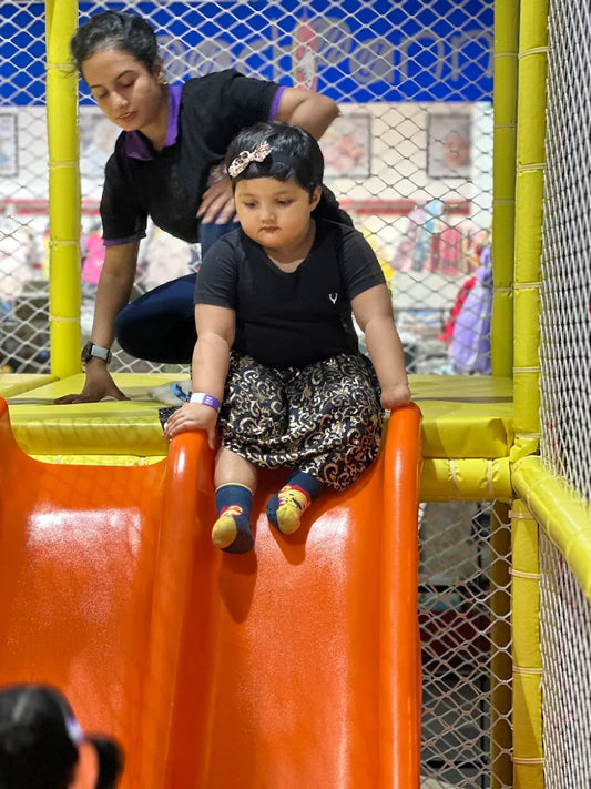 A young child sits on an orange slide in a play area, with an adult nearby.