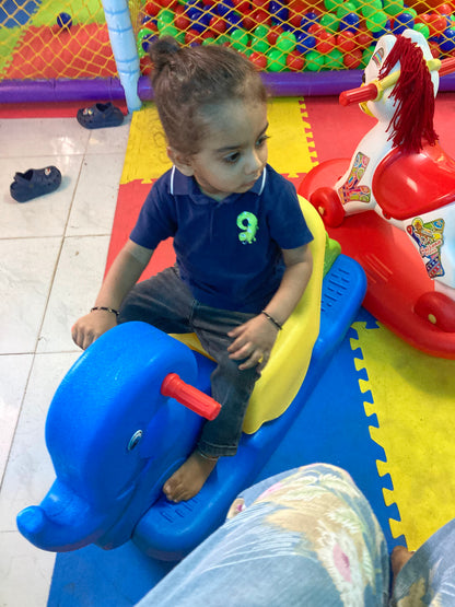 A young child rides a blue rocking toy in a play area with a ball pit in the background.