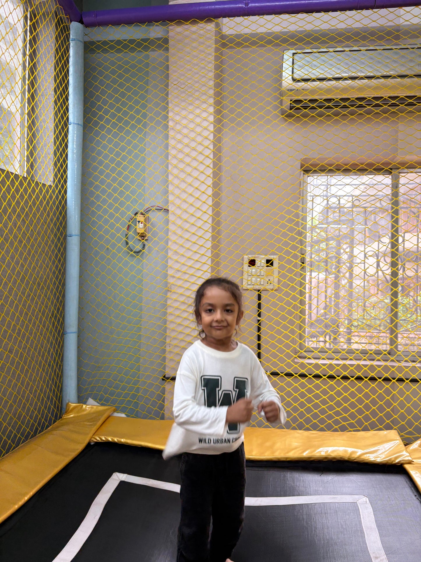 A young child jumps on a trampoline inside a play area with yellow netting.