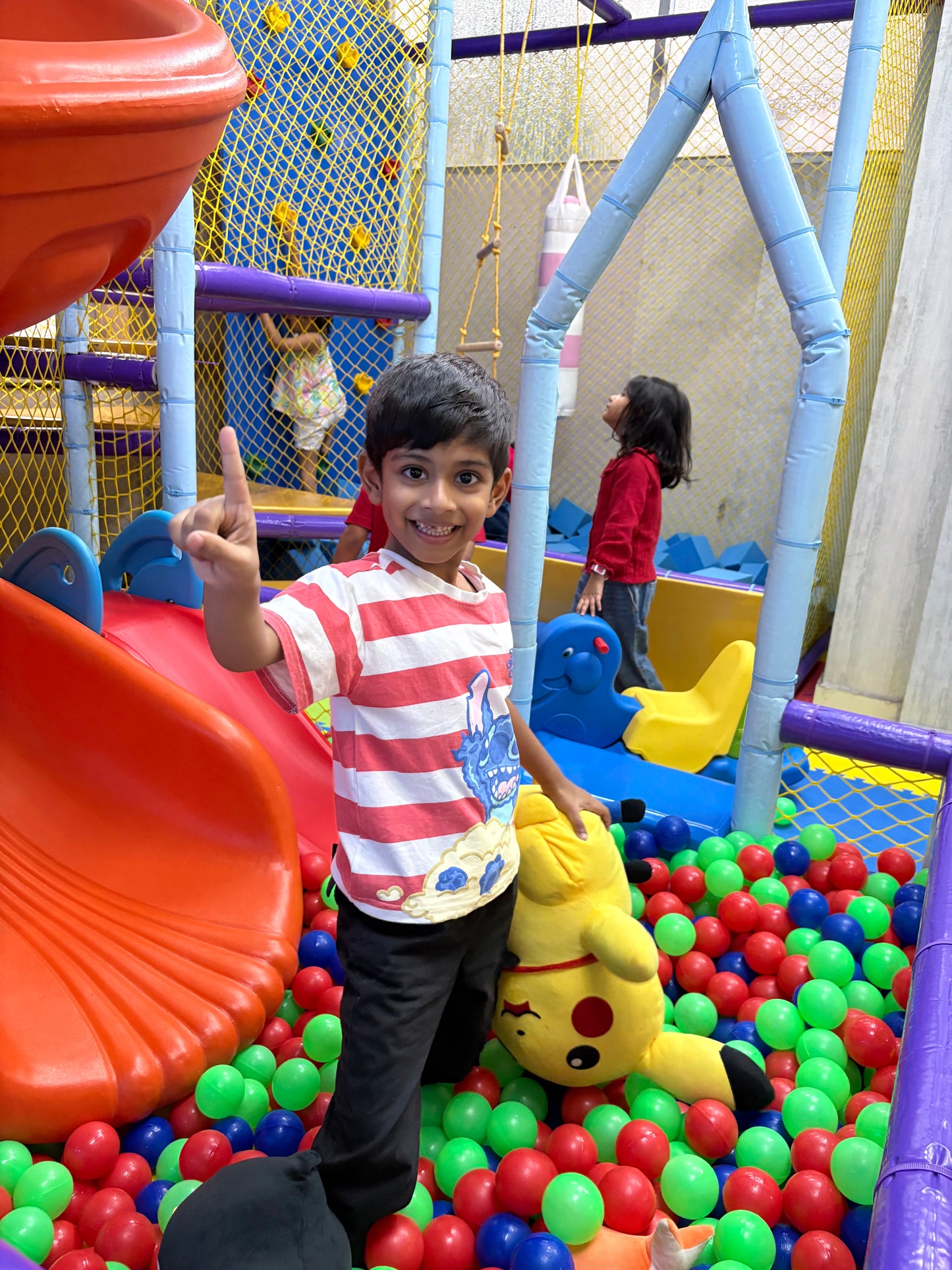 A young boy smiles and points up in a colorful indoor playground filled with ball pits and slides.