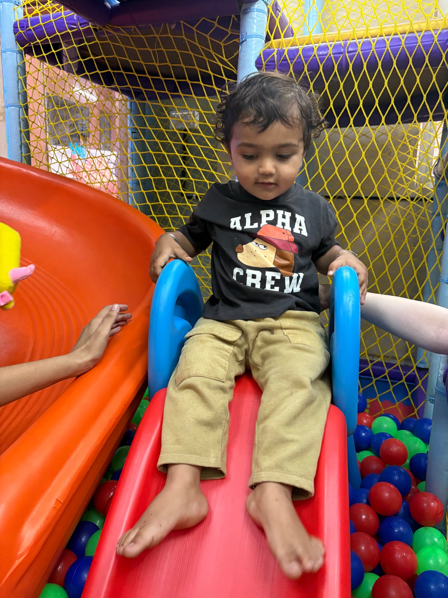 A young child sits on a slide in a ball pit, wearing a t-shirt with a dog graphic.
