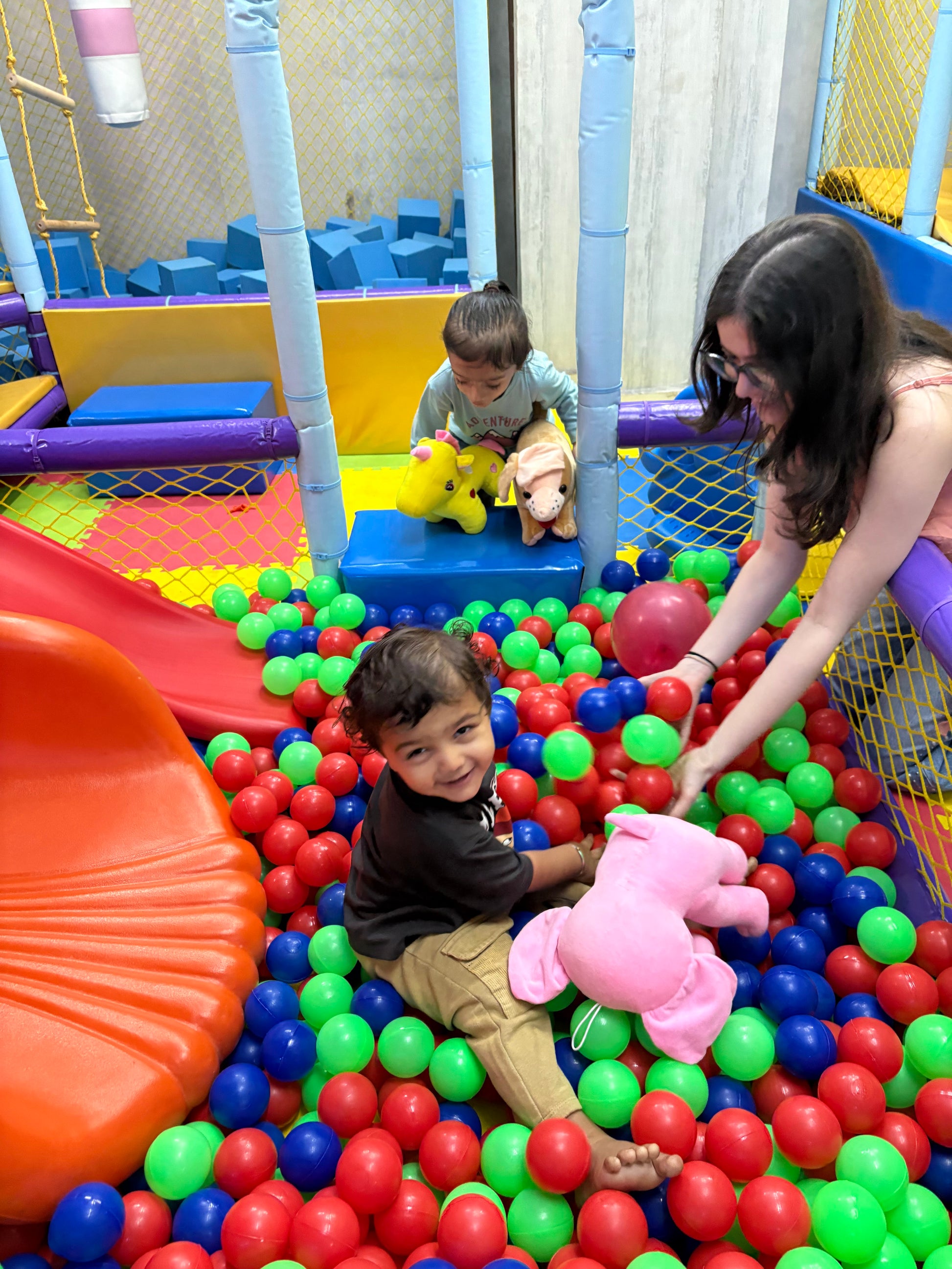 A young child smiles while playing in a ball pit with a woman and another child in the background.
