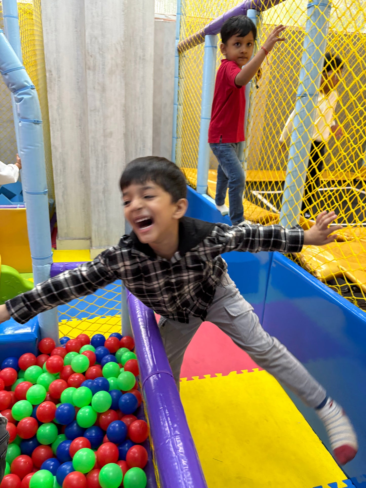 A young boy laughs joyfully as he plays in a colorful ball pit at an indoor playground. Another child is visible in the background.
