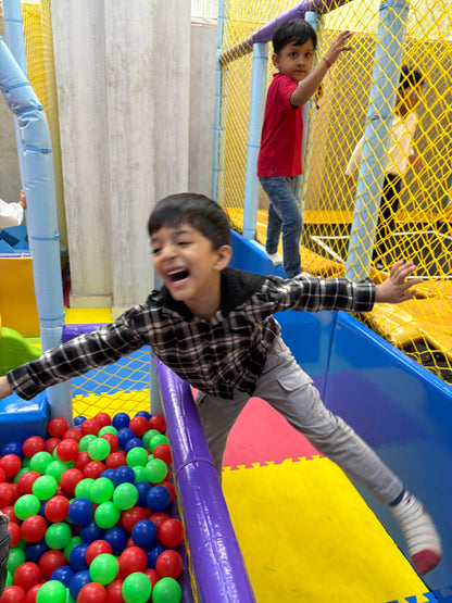 A young boy laughs joyfully as he plays in a colorful ball pit at an indoor playground. Another child is visible in the background.