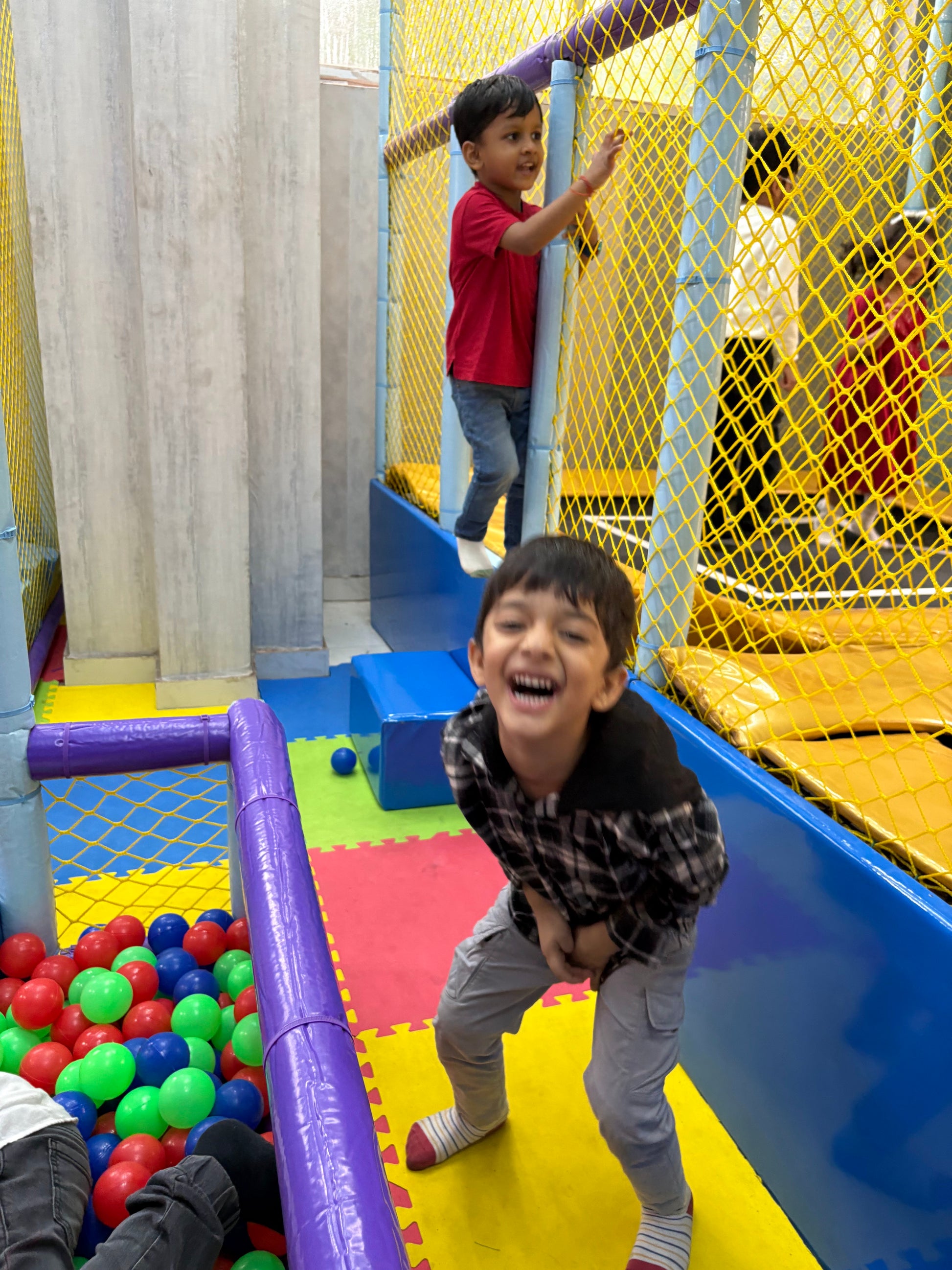 Two young boys play in a colorful indoor playground with a ball pit. One boy laughs joyfully while the other stands on a platform.