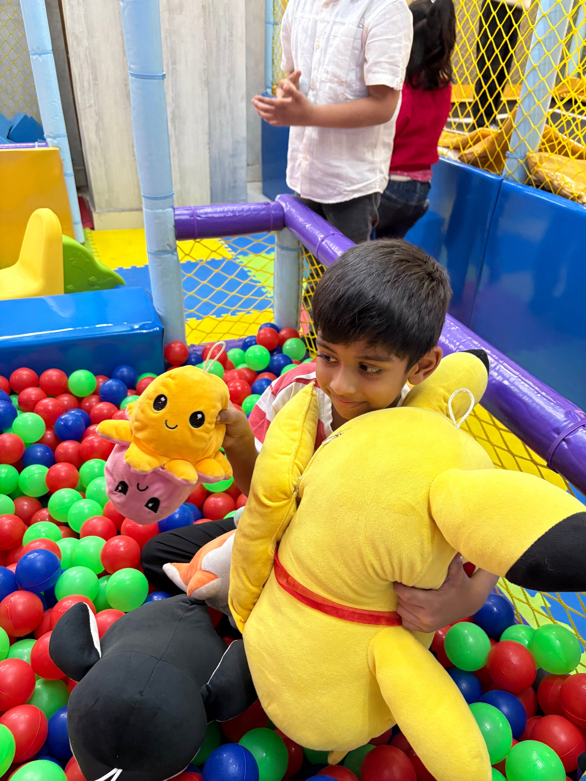 A young boy in a ball pit holds stuffed toys, with other children in the background.