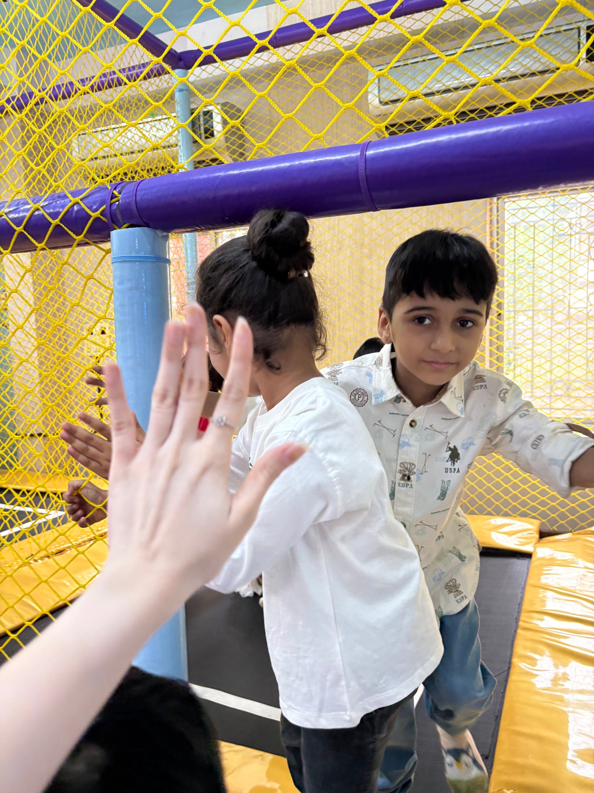 Two children play in a colorful indoor playground. A hand is held up in the foreground.
