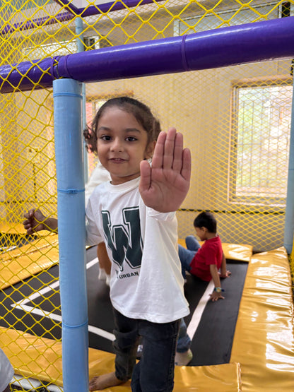 A young child holds up their hand in a play area with trampolines. Another child sits on a trampoline in the background.