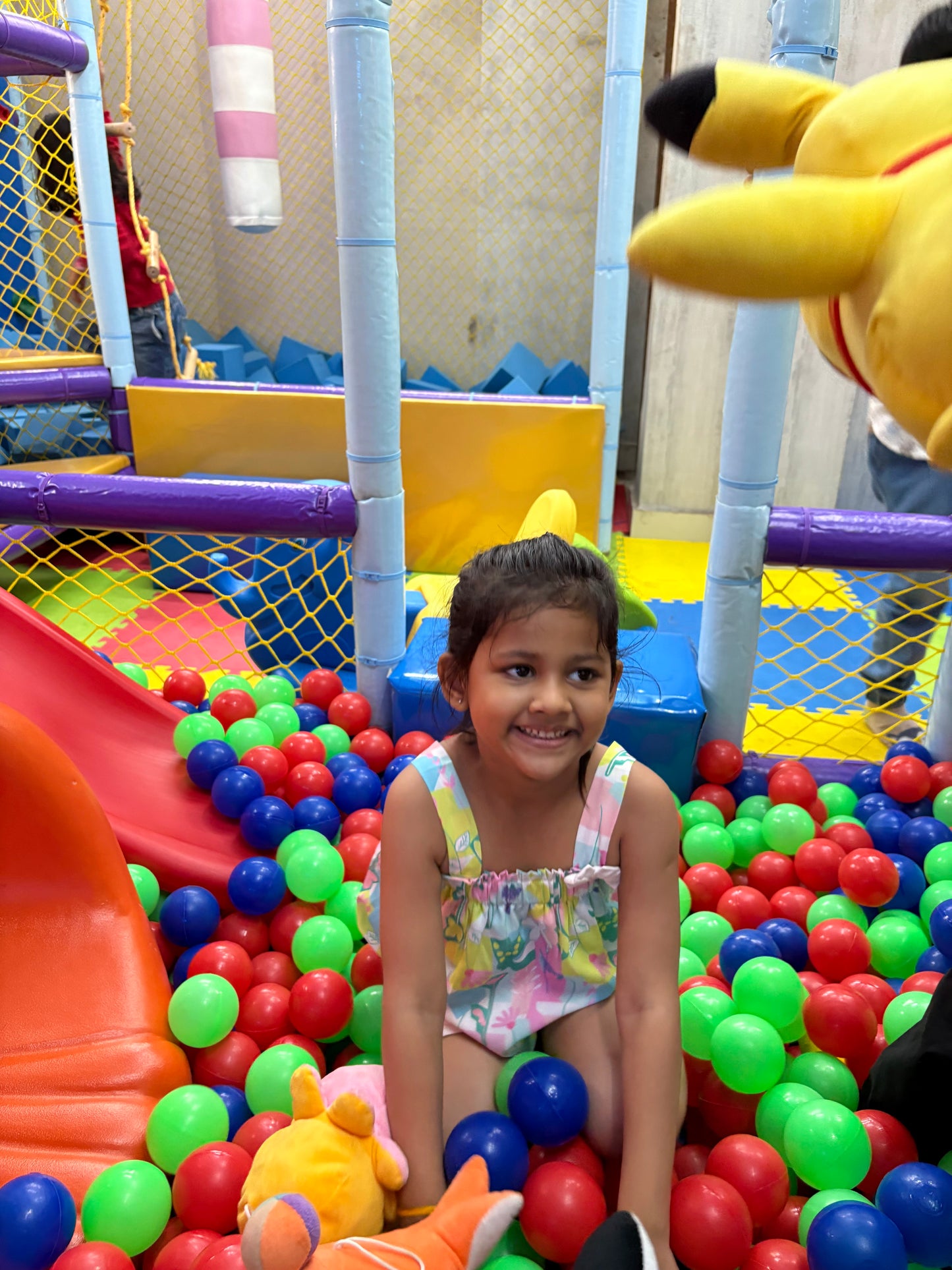 A young girl smiles in a ball pit with a red slide and colorful balls.